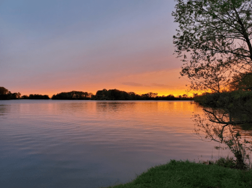 A serene lake at sunset, with vibrant orange and purple hues reflecting on the water, surrounded by trees.