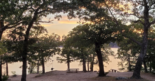 A serene lakeside view at sunset, framed by trees with picnic tables in the foreground.