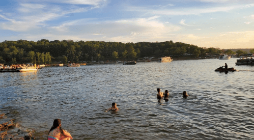 People swimming and relaxing by a lake during sunset, with boats and trees in the background.