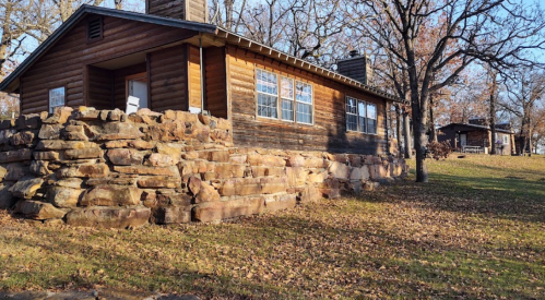 A rustic wooden cabin with a stone foundation, surrounded by trees and fallen leaves in a sunny outdoor setting.