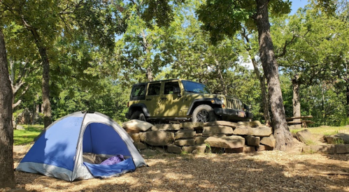 A blue tent is set up on a rocky area, with a green Jeep parked nearby under trees in a sunny outdoor setting.