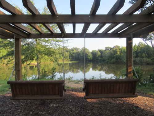 Two wooden swings hang from a pergola, overlooking a serene lake surrounded by trees.