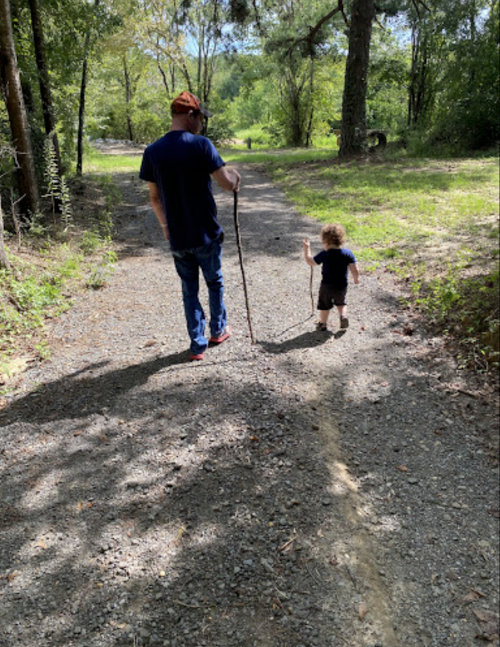 A man walks on a gravel path with a stick, while a small child crawls beside him in a green, wooded area.