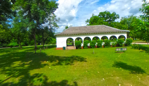A white pavilion with arches surrounded by greenery and trees under a partly cloudy sky.