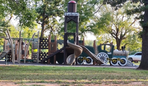 Playground featuring a train-themed structure with slides and climbing areas, surrounded by trees and grass.