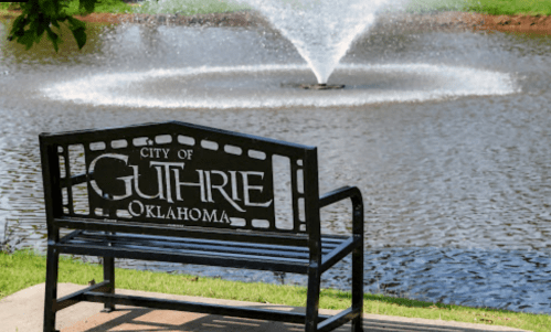 A black bench with "City of Guthrie, Oklahoma" engraved, overlooking a pond with a fountain.
