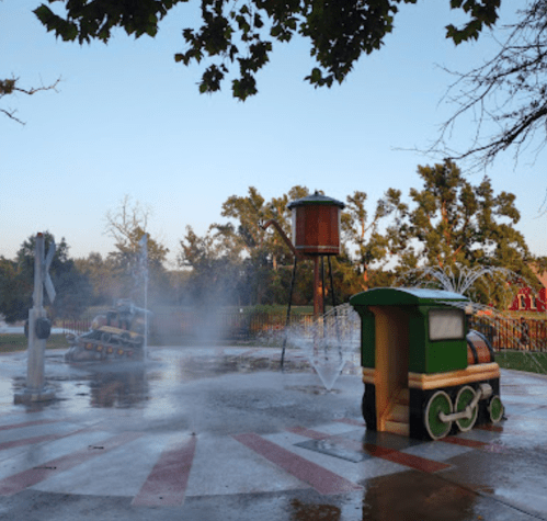 A colorful splash pad with water features, including a train and a water tower, surrounded by trees and a clear sky.