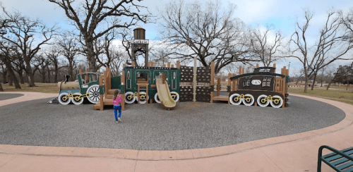 A colorful playground resembling a train, with slides and climbing structures, set in a park with bare trees.