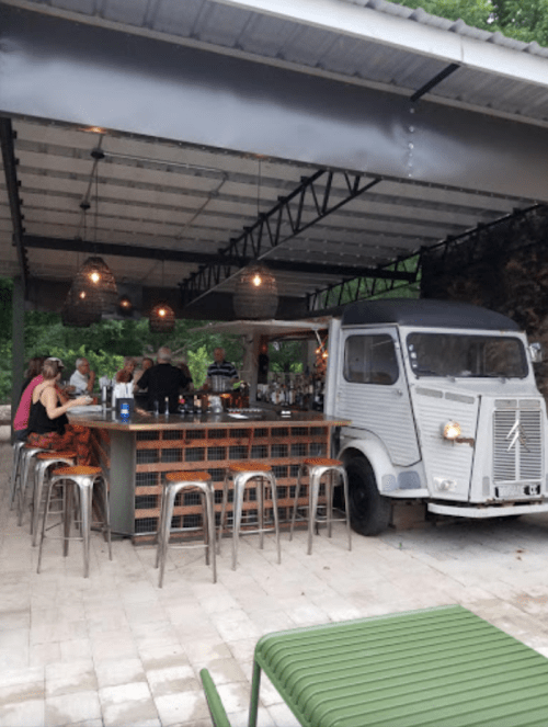 A rustic outdoor bar with a vintage truck, surrounded by patrons enjoying drinks under hanging lights.
