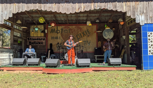 A band performs on stage at a rustic venue, with colorful decor and instruments set up in front of them.