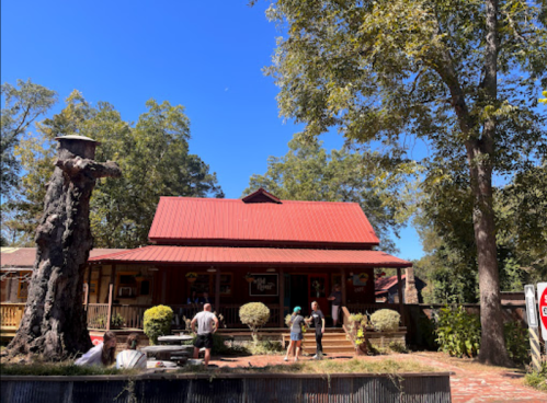 A rustic building with a red roof surrounded by trees, featuring people gathered outside on a sunny day.