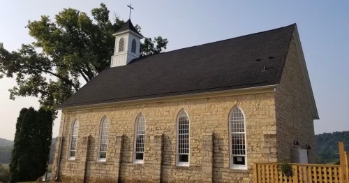 A stone church with arched windows and a bell tower, set against a clear sky and green trees.