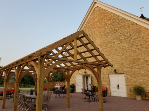 A wooden pergola in front of a stone building with outdoor seating and potted flowers.