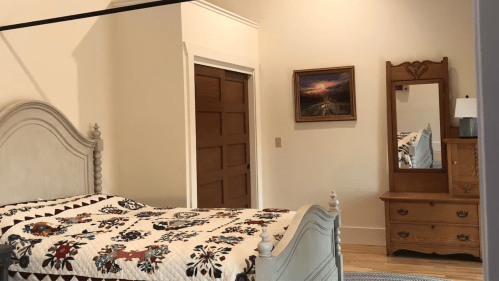 A cozy bedroom featuring a quilted bed, wooden dresser, mirror, and a landscape painting on the wall.