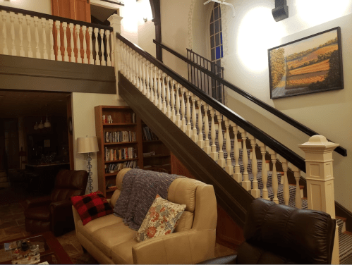 A cozy living room with a staircase, bookshelves, and a painting on the wall, featuring comfortable seating.