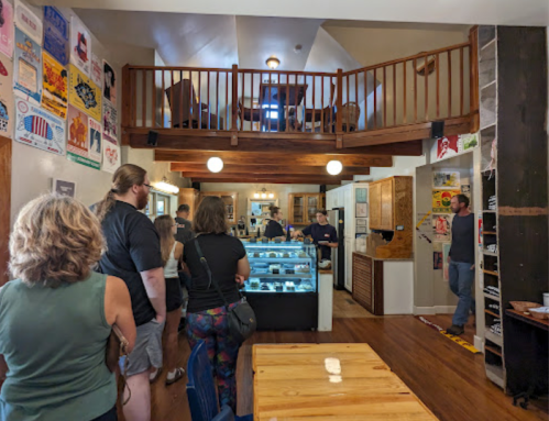 A cozy café interior with a counter, customers in line, and a second floor with seating and posters on the walls.