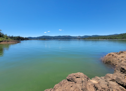 A serene lake surrounded by green hills under a clear blue sky, with rocky shores in the foreground.