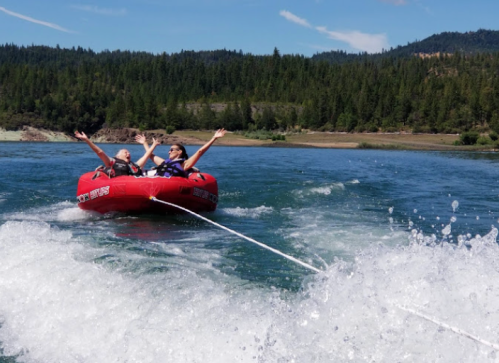 Two people joyfully tubing on a lake, arms raised, with a scenic forest backdrop and splashing water.