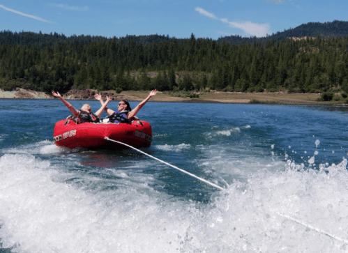 Two people joyfully tubing on a lake, arms raised, with a scenic forest backdrop and splashing water.