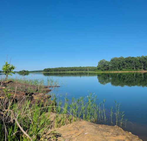 A calm lake reflects a clear blue sky, surrounded by green trees and rocky shoreline.