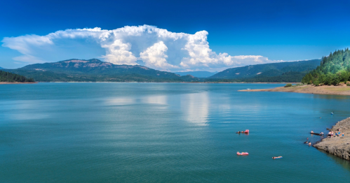 A serene lake surrounded by mountains, with fluffy clouds in a blue sky and people enjoying the water.