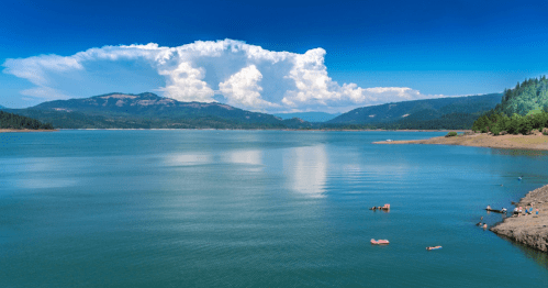 A serene lake surrounded by mountains, with fluffy clouds in a blue sky and people enjoying the water.