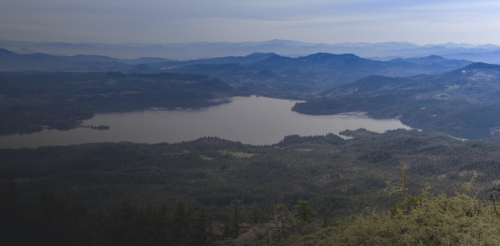 A panoramic view of a serene lake surrounded by mountains and forests under a clear blue sky.