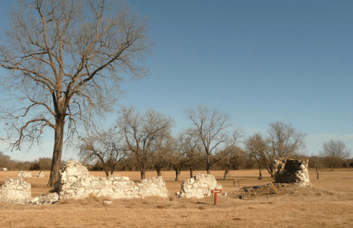 Ruins of a stone structure surrounded by bare trees and open fields under a clear blue sky.