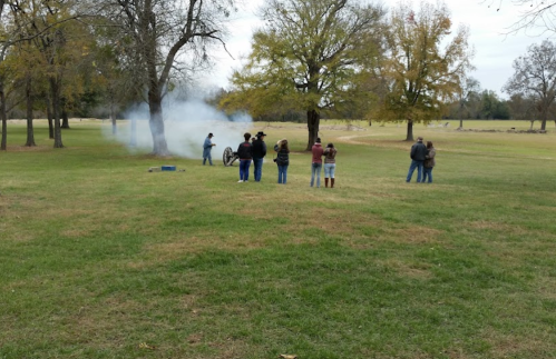 A group of people stands in a grassy field, watching smoke rise from a cannon being fired nearby.