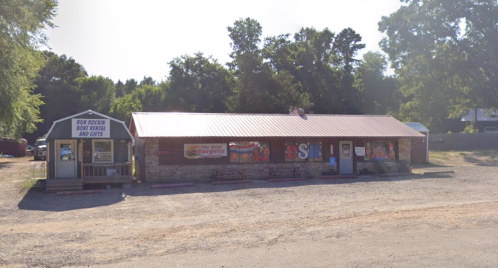 A rustic building with a metal roof, featuring signs for a bait shop and gifts, surrounded by trees and gravel.