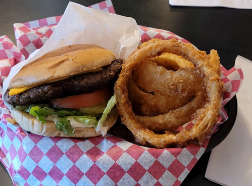 A cheeseburger with lettuce and tomato, served with crispy onion rings in a red and white checkered basket.