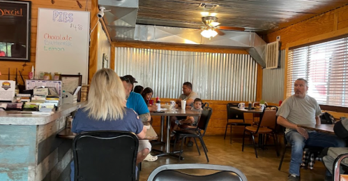 A cozy diner interior with patrons seated at tables, wooden decor, and a menu board displaying pie flavors.