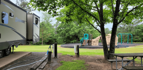 A camper parked near a playground with a sand area and slides, surrounded by green trees and grass.