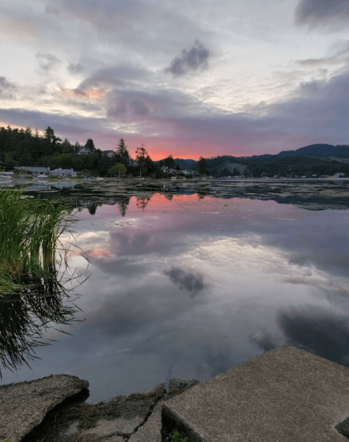 A serene lake at sunset, reflecting colorful clouds and trees, with a rocky shore in the foreground.