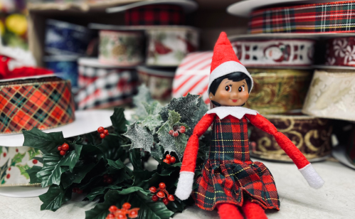 A festive elf doll in a plaid outfit sits among colorful ribbons and holly decorations.