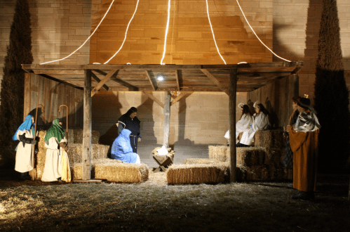 A nativity scene with figures in traditional attire gathered around a manger under a lit canopy. Hay bales surround them.