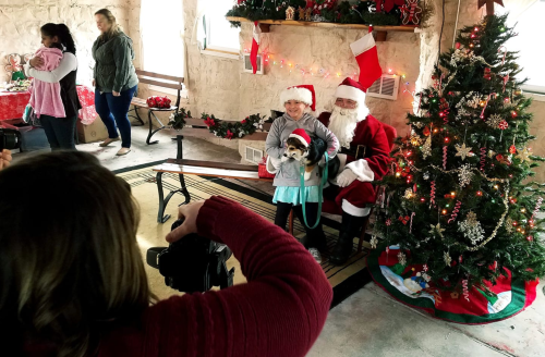 A child in a Santa hat poses with Santa Claus in front of a decorated Christmas tree, while someone takes a photo.