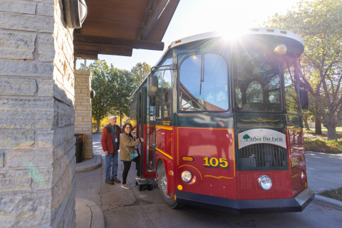 A red trolley bus parked outside a building, with two people standing nearby, enjoying a sunny day.