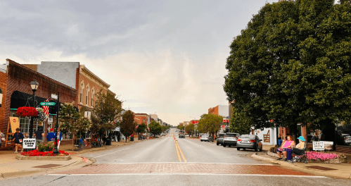 A quiet downtown street lined with shops, trees, and parked cars under a cloudy sky. People sit on benches.