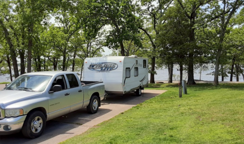 A silver pickup truck towing a white travel trailer near a lake, surrounded by green trees and grass.