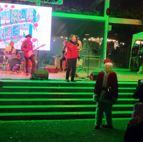 A festive outdoor concert scene with a band performing on stage and a person in a Santa costume in the foreground.
