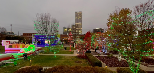 A festive park scene with colorful holiday lights on trees and decorations, set against a city skyline.