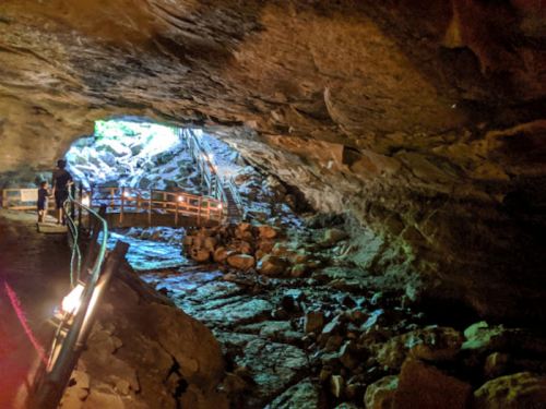 A cave entrance with a rocky floor and wooden walkway, illuminated by natural light from outside.