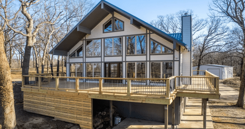 A modern cabin with large windows and a wooden deck, surrounded by trees on a clear day.