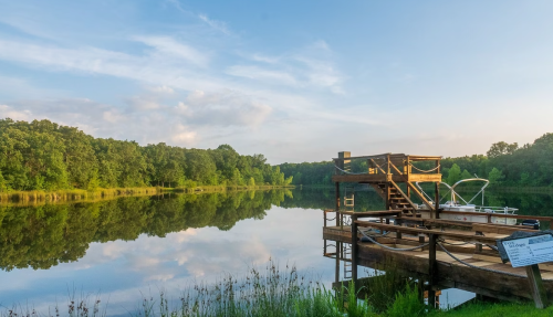 A serene lake scene with a wooden dock, surrounded by lush greenery and reflecting clouds in the calm water.