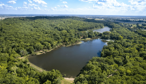 Aerial view of a serene lake surrounded by lush green trees and rolling hills under a blue sky with scattered clouds.