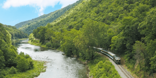 A train winds along a riverbank, surrounded by lush green hills and trees under a blue sky.