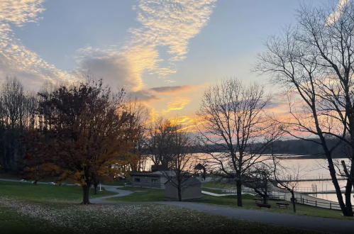 A serene lakeside view at sunset, with trees, clouds, and a small building in the foreground.