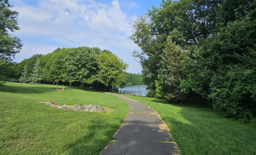 A winding path through lush green grass and trees, leading to a serene lake under a partly cloudy sky.