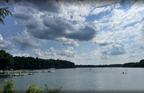 A serene lake scene with boats, lush greenery, and a cloudy sky reflecting on the water.